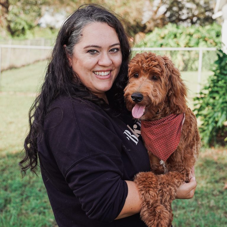 Woman with black hair wearing a black shirt holding a red goldendoodle dog who is wearing a red bandana
